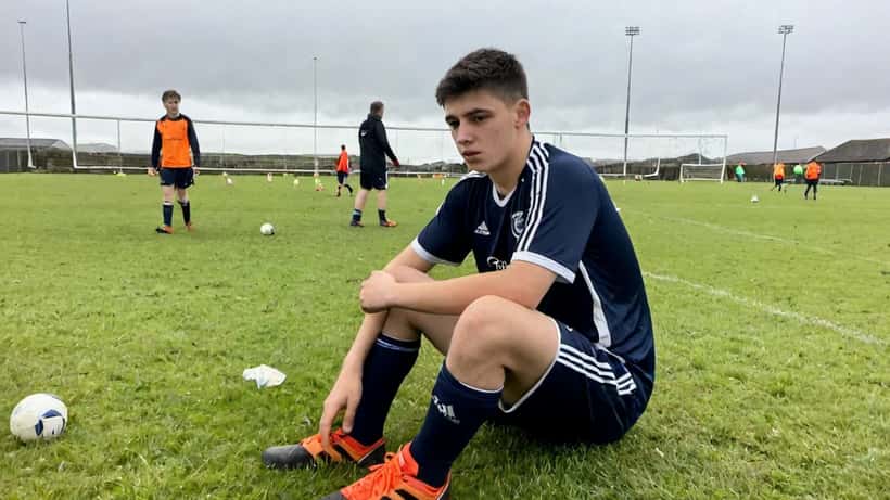 Young academy footballer sitting on training pitch holding his knee after injury, coach beside him, grey overcast sky