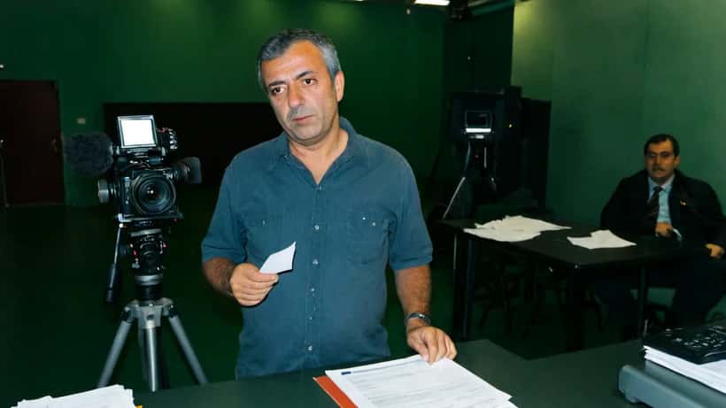 Apresentador de televisão diante de estúdio de TV português com documentos de contrato laboral na mesa